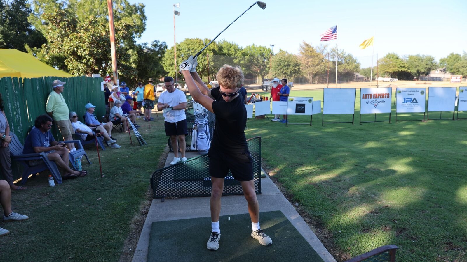 A golfer in black at the top of the backswing on the tee at the 2026 Hole-in-One Shootout, with sponsor signs lining the range