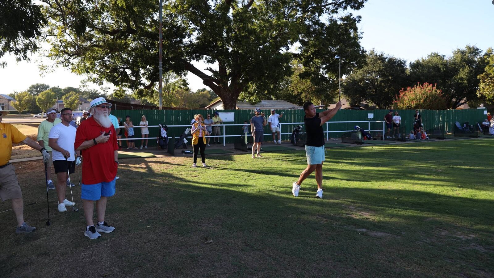 Players and spectators, including a man with a white beard in a red shirt, watch a golfer follow through at the 2026 Hole-in-One Shootout