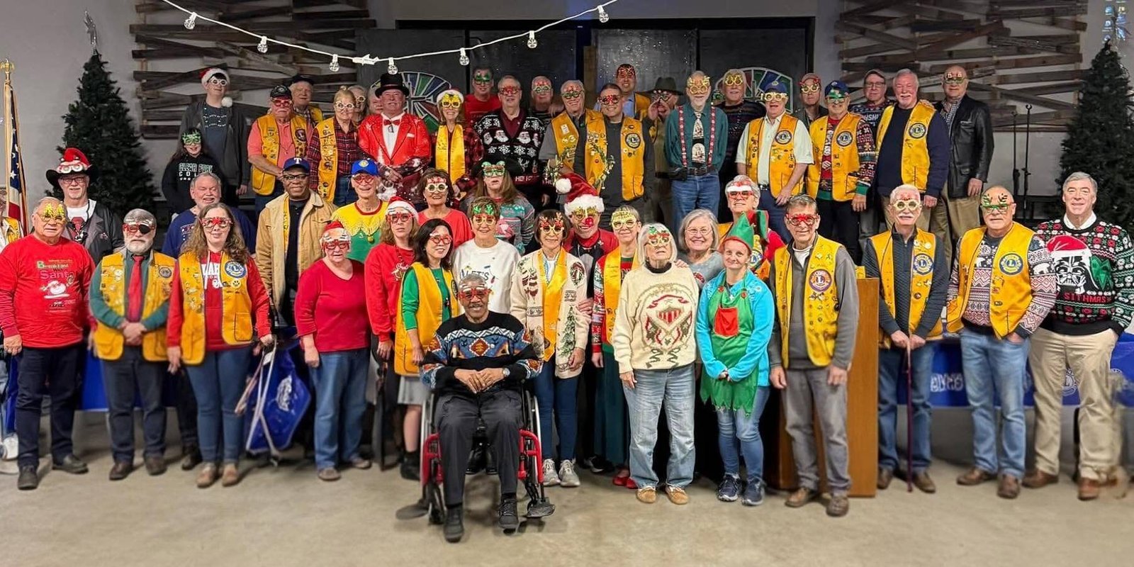 Dozens of Colleyville Lions in gold vests, holiday sweaters and festive glasses pose together for a club group photo under strings of lights
